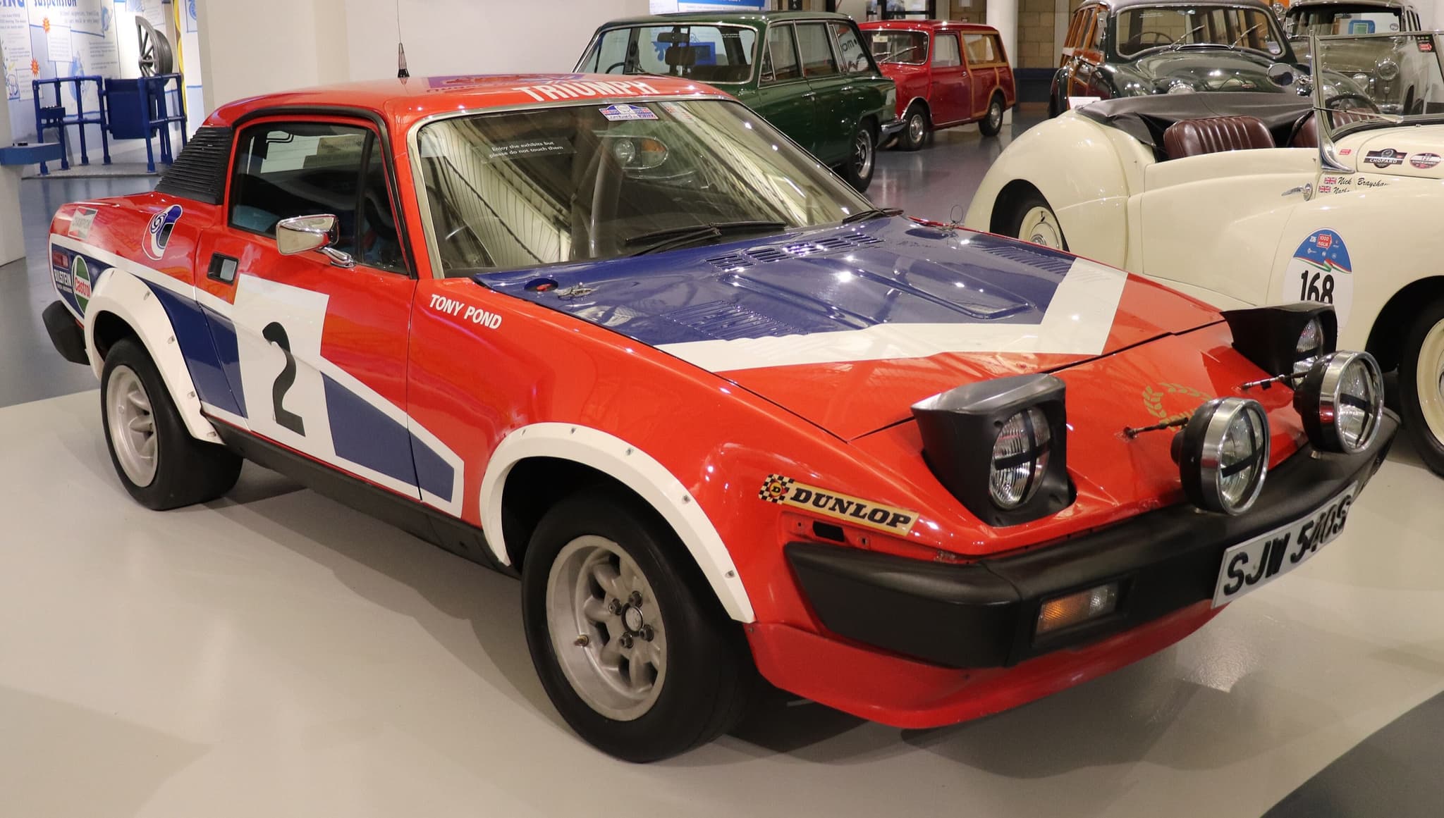A red, white and blue TR7 V8 rally car, number 2, with four lamps in its nose, on a museum floor.