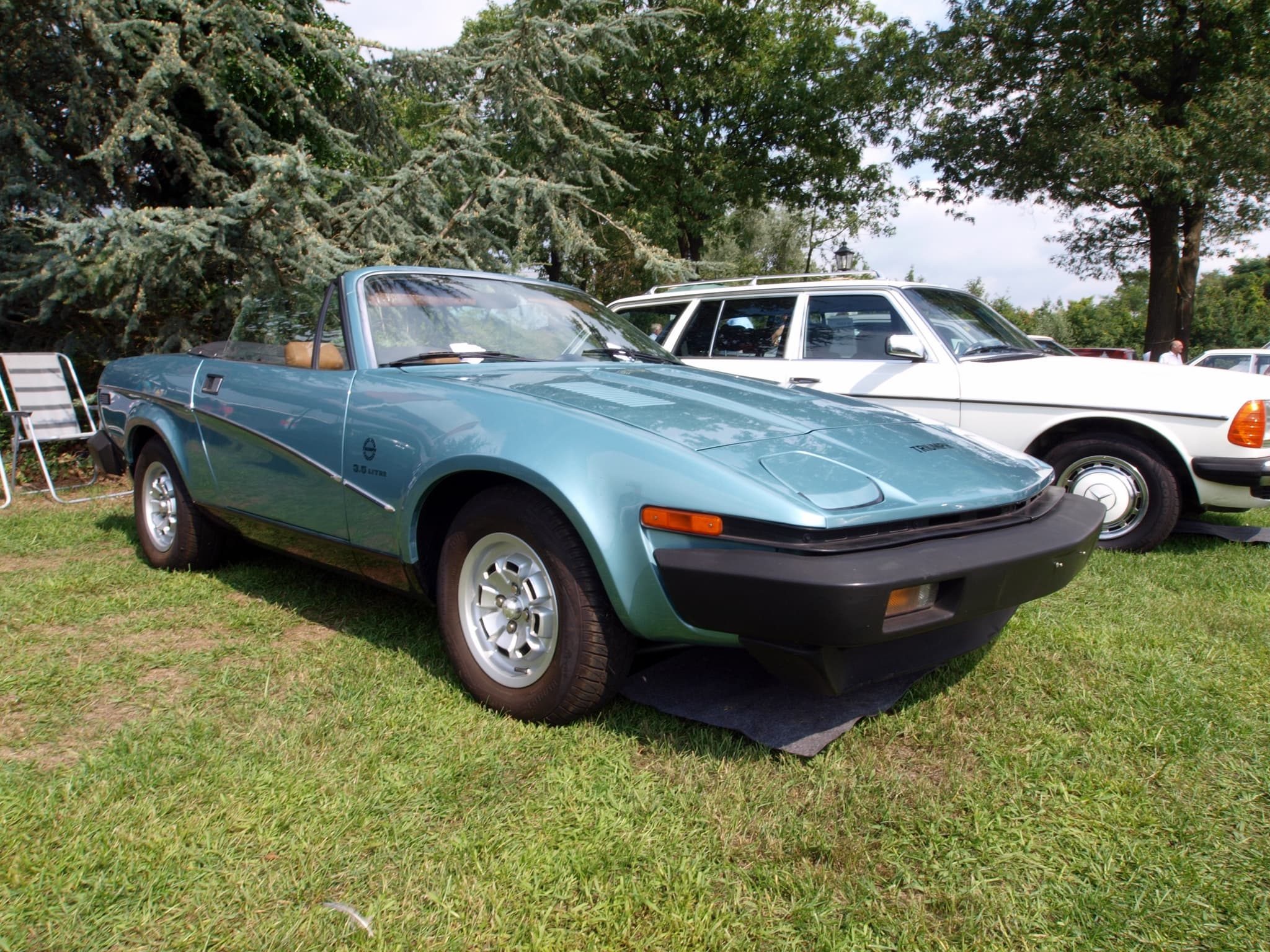 A pale blue wedge-shaped convertible, its top down, parked on grass beside a white estate car under tall trees.