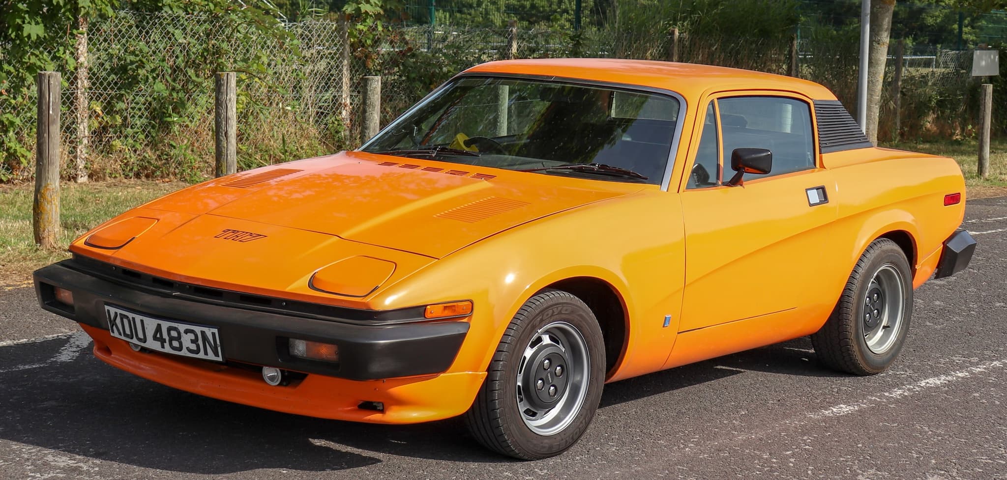 An orange TR7 coupé with steel wheels, front three-quarter in a sunny car park by a chain-link fence.
