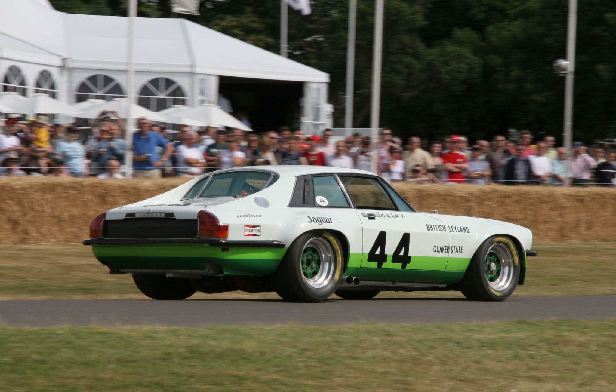 A white and green Jaguar XJ-S racing car, number 44, running uphill past straw bales and a crowd.