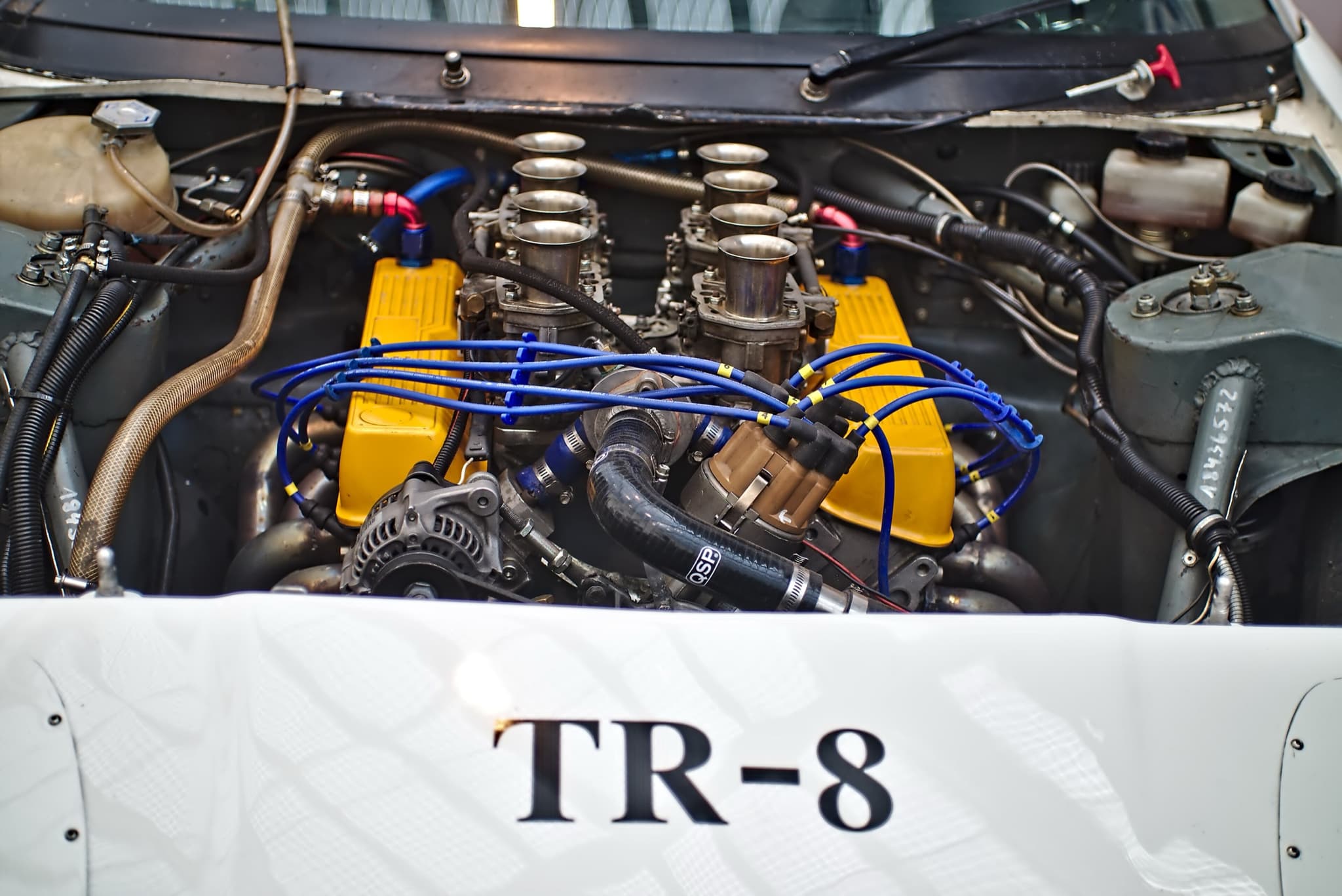 A racing V8 with yellow rocker covers and eight polished carburettor trumpets, TR-8 lettered on the panel in front of it.