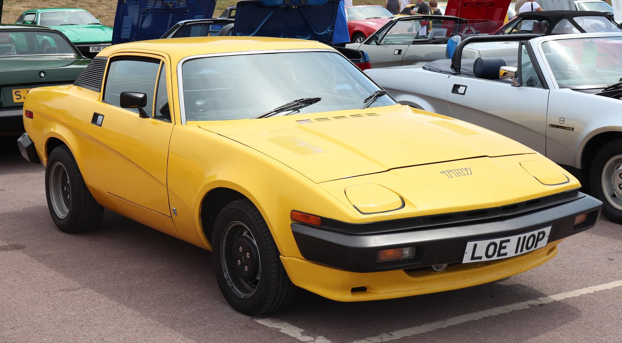 A yellow TR7 coupé with black steel wheels in a show car park, a silver wedge convertible beside it.