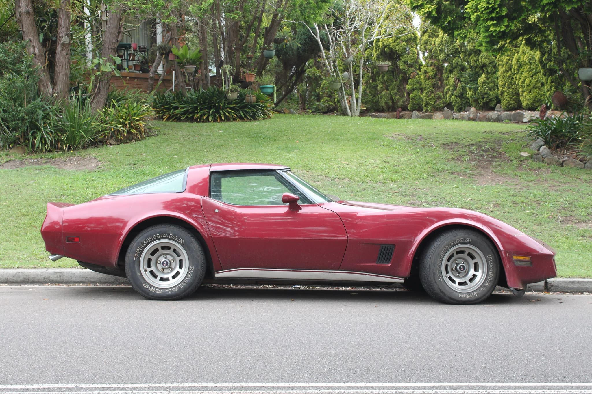 A maroon Chevrolet Corvette coupé in profile, parked at a kerb in front of a grassy bank.