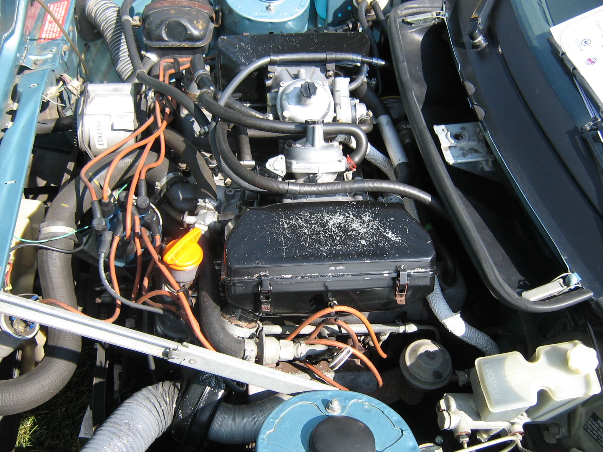The engine bay of a light blue TR8, the V8 fed by a carburettor under a black airbox, with orange ignition leads.