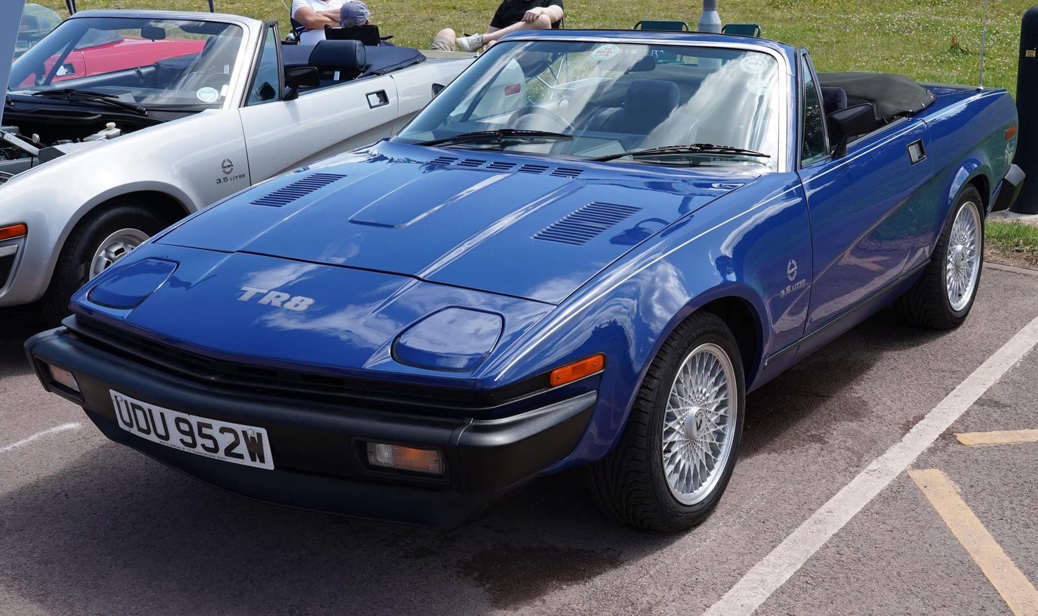 A bright blue wedge convertible with TR8 decals, its hood folded and cross-spoke alloy wheels, in a show car park.