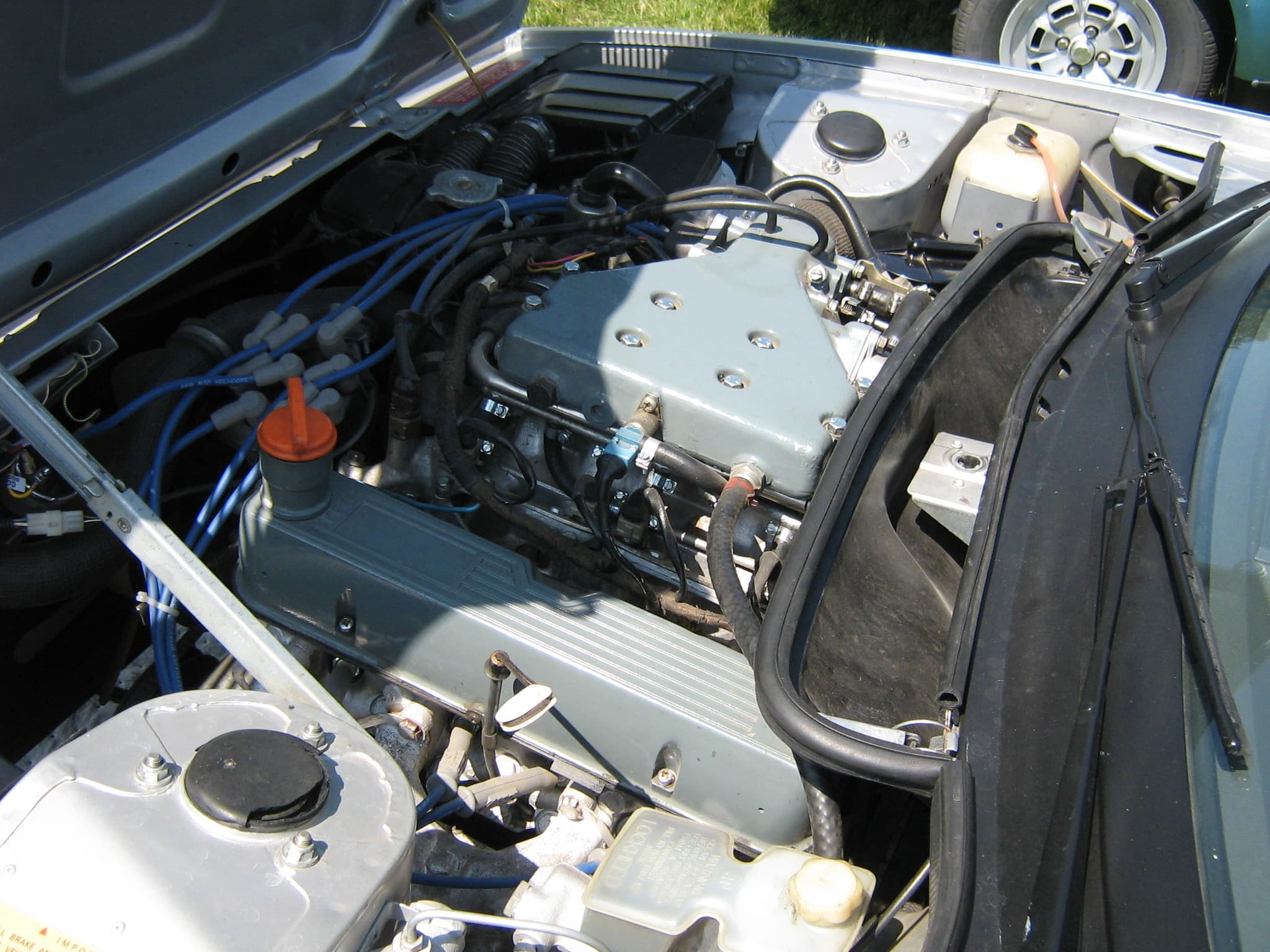 The engine bay of a silver TR8, the V8 topped by the grey cast plenum of its fuel injection, with blue ignition leads.