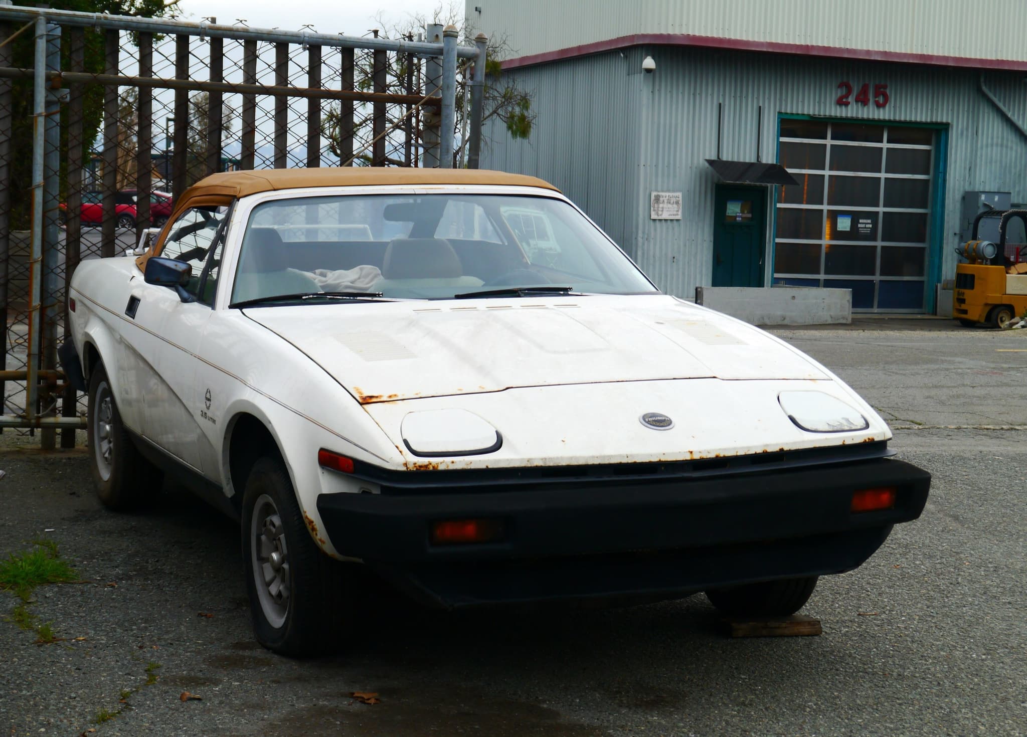 A white wedge convertible with a tan hood and rust along its bonnet edge and front wing, in an industrial yard.