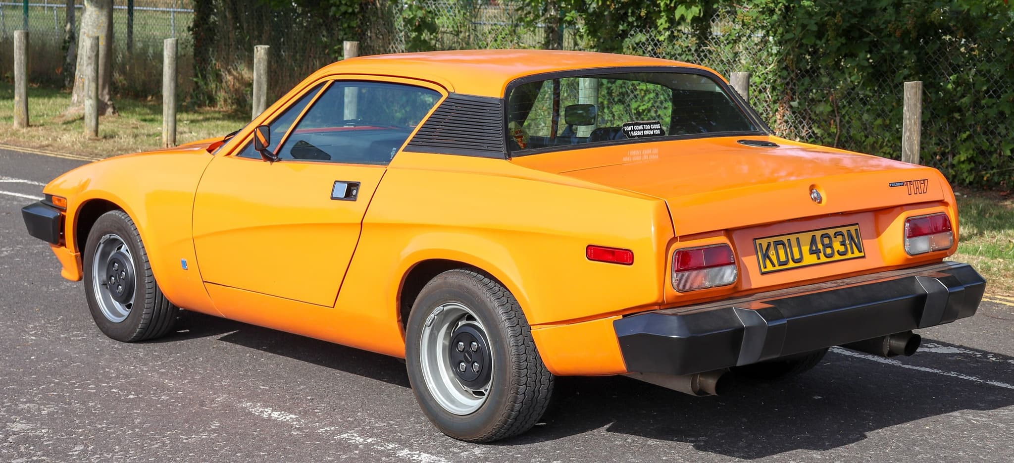 An orange TR7 coupé from behind, with TRIUMPH and TR7 badges on the tail and a tailpipe at each side.