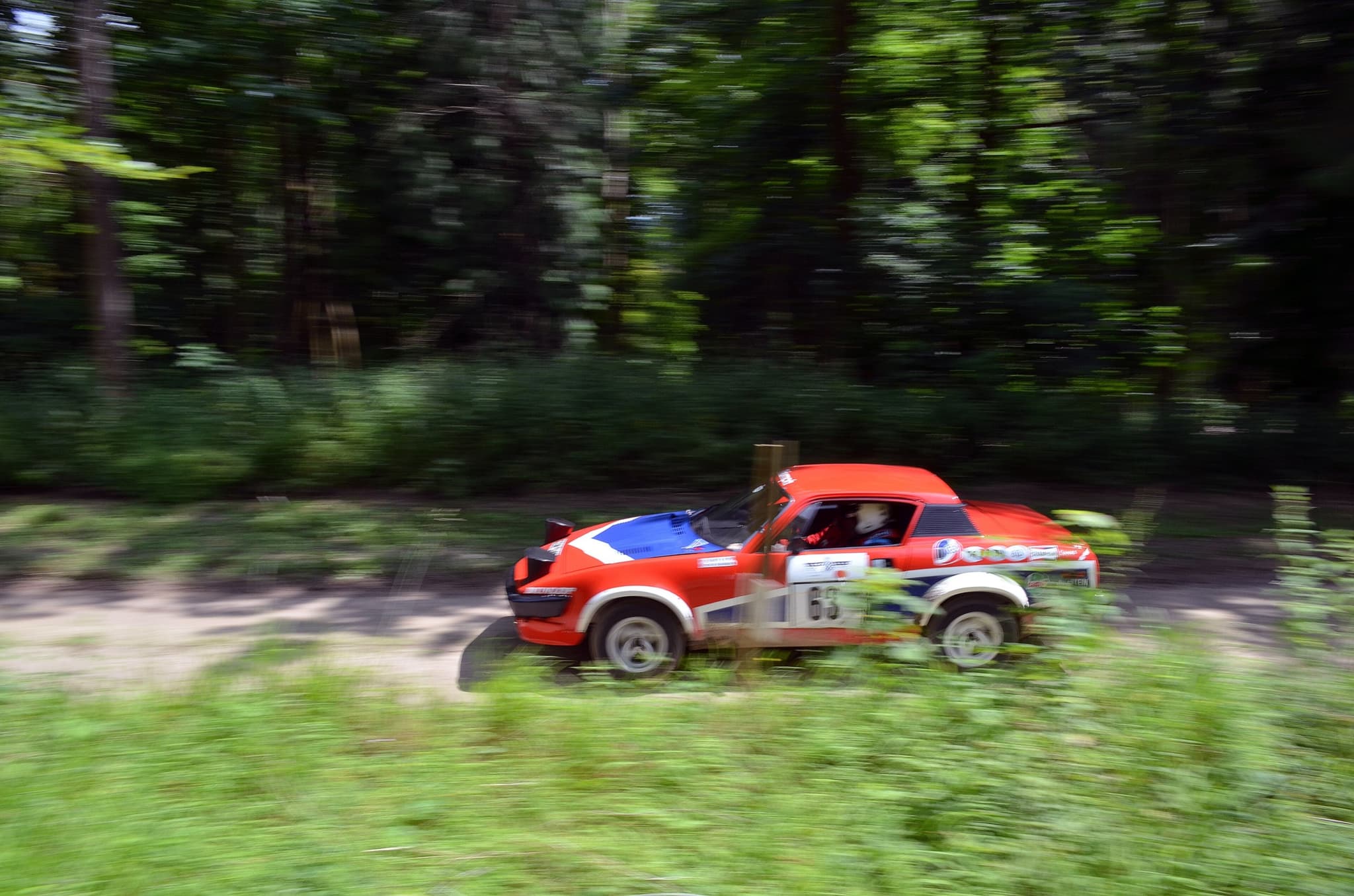 A red, white and blue TR7 V8 rally car, number 63, at speed on a sandy forest track. Opens a larger view
