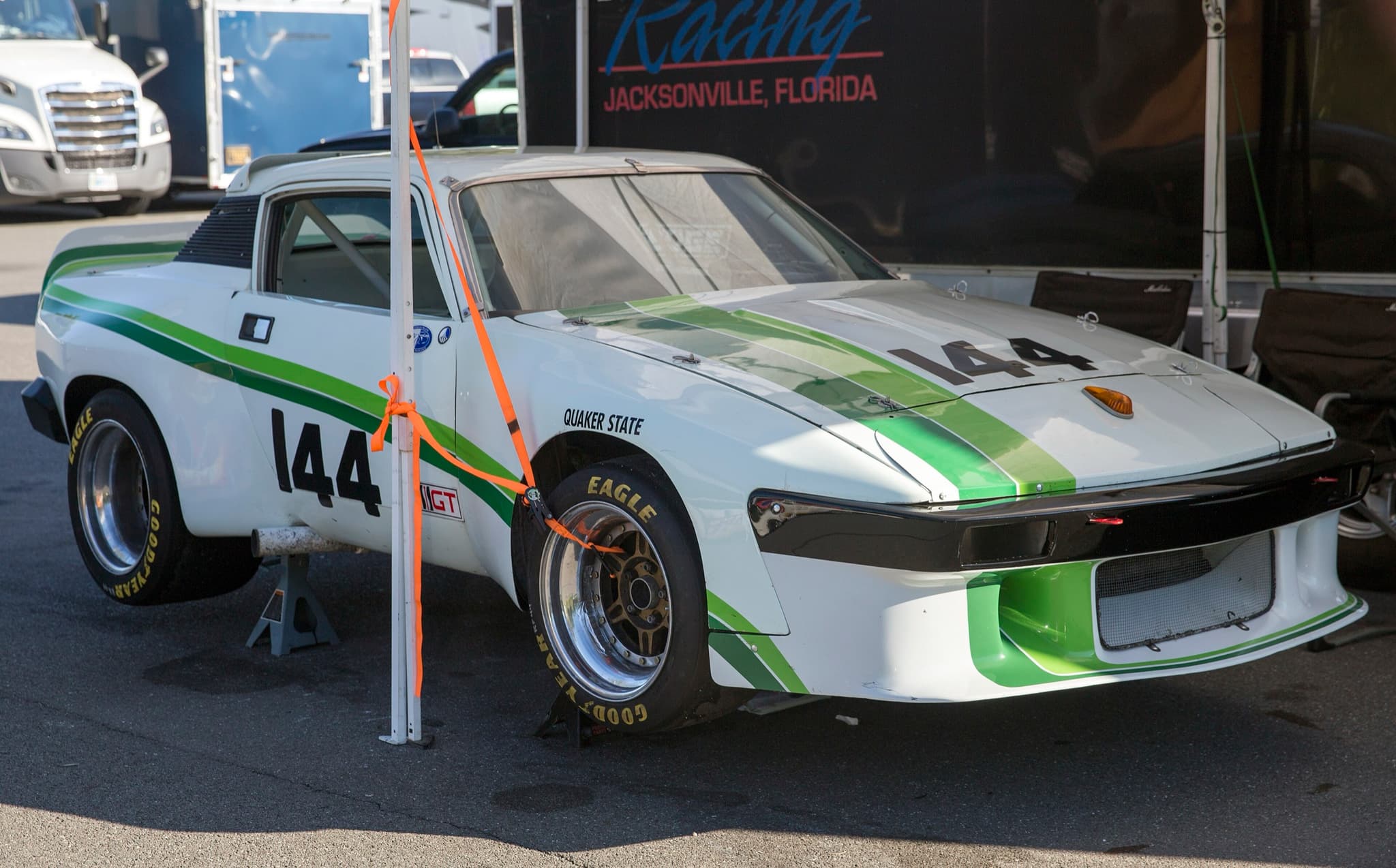 A white racing TR8 coupé with green stripes and wide arches, number 144, on axle stands under a paddock gazebo.
